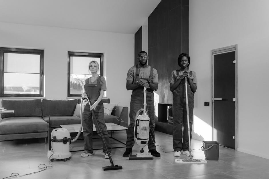 A professional cleaning team at work in a modern kitchen, featuring three staff members wearing uniforms and using cleaning tools. One person stands on a step stool, cleaning the upper kitchen cabinet