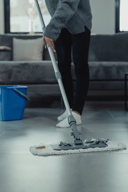 Close-up view of three professional cleaning workers in gray t-shirts and bright orange overalls, standing indoors on a tiled floor, holding vacuum cleaners with their hands. The image showcases the e