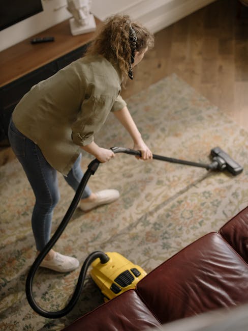 A woman is engaged in domestic cleaning, vacuuming an intricate patterned area rug with a yellow and black upright vacuum cleaner in a well-lit living room. She is wearing a khaki jacket, blue jeans, and white sneakers, with curly hair tied back. The wooden flooring surrounding the rug is visible, along with a dark wood sideboard against the wall holding a small decorative statue. The scene illustrates surface cleaning and deep cleaning practices, emphasizing hygiene and upkeep in a residential setting. Maida Vale Cleaner specializes in detailed, professional cleaning services for homes, including carpet maintenance and sanitisation, as highlighted in the Little Venice carpet cleaning insider tips blog.