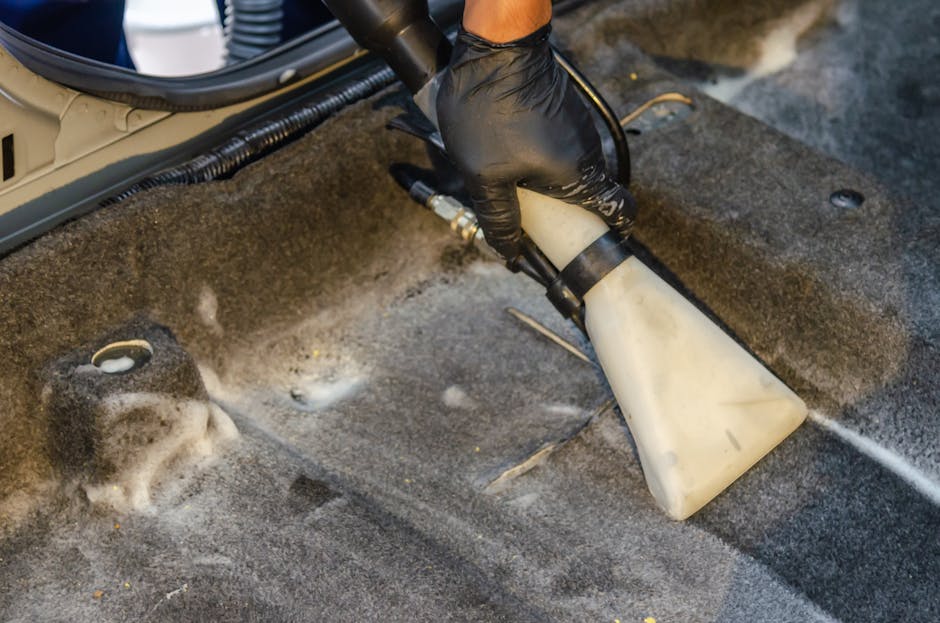 A cleaning technician wearing black gloves is using a handheld steam or spray device to deep clean and sanitize the carpeted floor of a residential or commercial space, with visible foam and water residue. The carpet appears to be a dark, textured material, possibly synthetic or wool, and is situated in a room with a neutral-colored floor. The device is directed at the carpet, indicating surface cleaning process. Nearby, there is a cylindrical object that may be part of the cleaning equipment, and the ambient lighting suggests a well-lit environment. The technician's hands and the cleaning tool are central in the image, highlighting professional carpet cleaning methods employed to maintain hygiene and surface cleanliness, consistent with services offered by Maida Vale Cleaner.