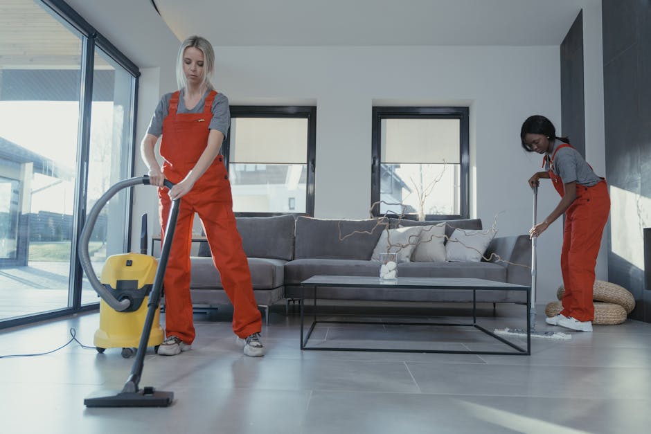 A young female cleaner with dark hair, dressed in a grey t-shirt and red overalls, holding a vacuum hose while standing in a well-lit modern living room with large windows. In the background, a male c
