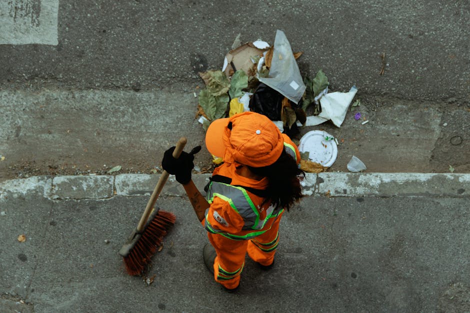 A street-level scene showing a sanitation worker in a bright green uniform and cap, wearing a dust mask, collecting debris from a large green wheeled trash bin placed on a paved sidewalk. The worker h